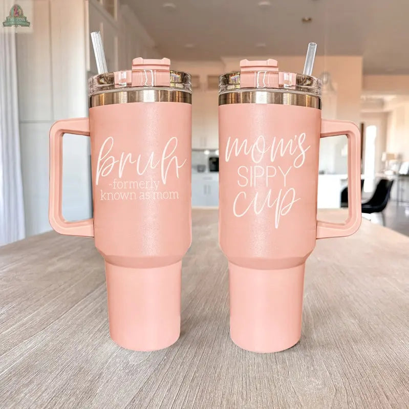 Two Bruh Pink 40oz tumblers with handles sit on a light wood table. The left one says "bruh - formerly known as mom," making it a perfect fun gift for moms. Both tumblers include clear lids and straws.