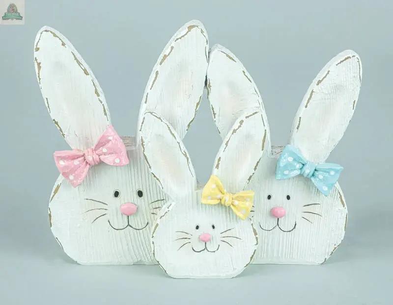 The Bunny Ears Family Tabletop features three wooden bunny figurines with white paint, pink noses, smiling faces, and polka-dotted bows in pink, yellow, or blue—charming spring décor for any space.