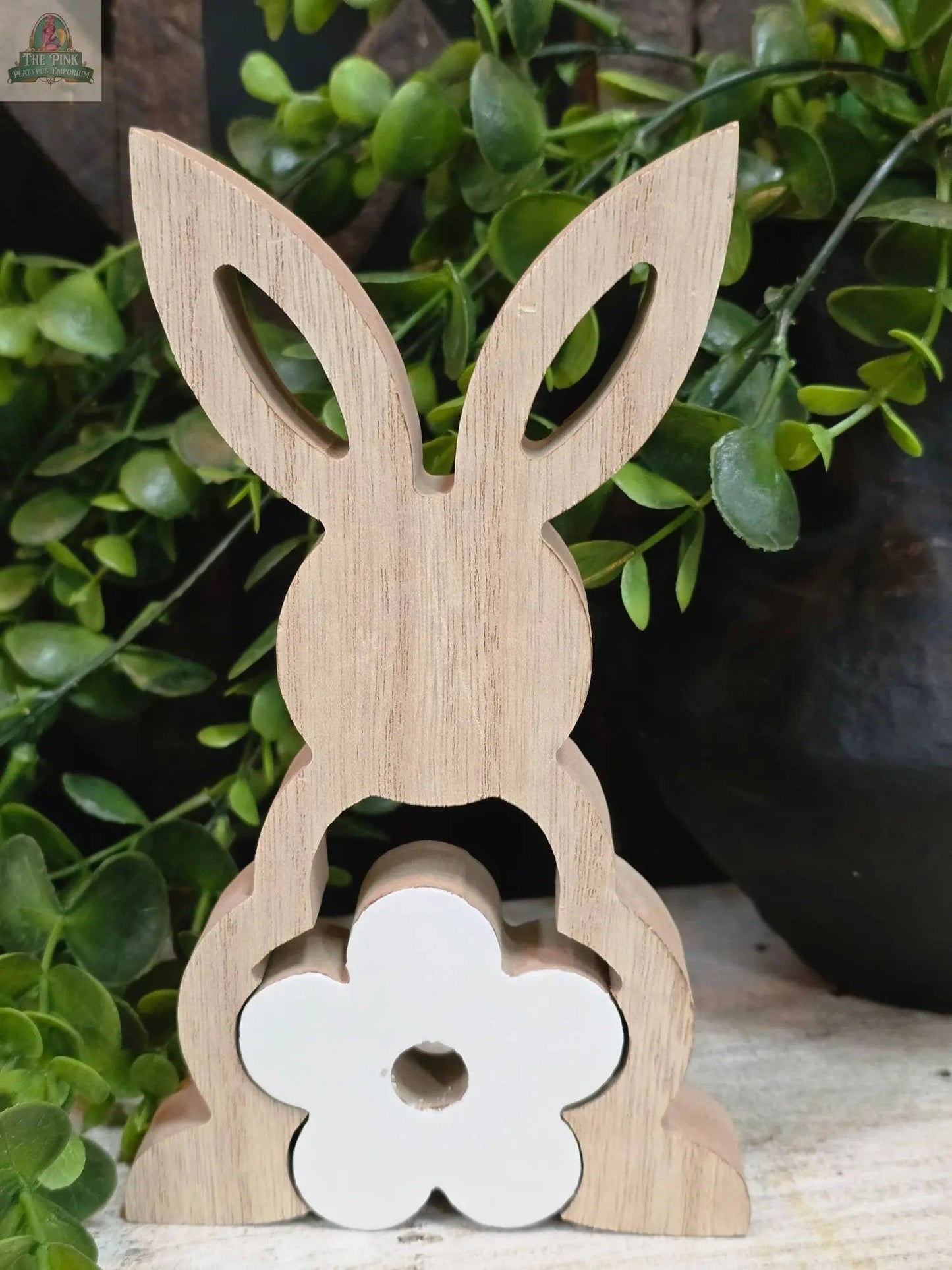 The Bunny with Enamel Flower Cutouts 3D 7in stands on a light surface, featuring 3D enamel flower details at its base, positioned in front of green leafy plants.