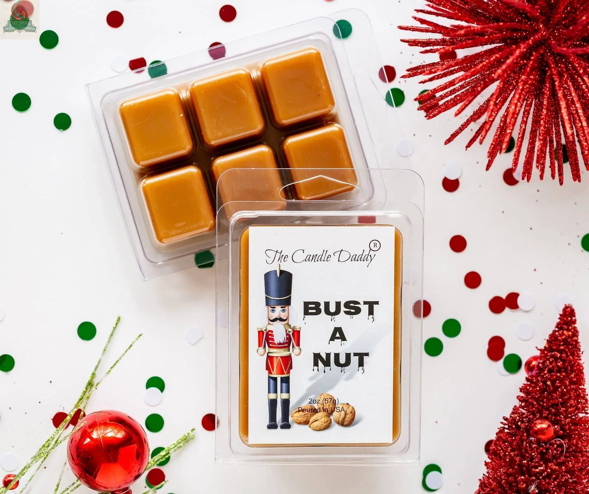 A pack of Candle Daddy’s “Bust A Nut – Funny Christmas Banana Nut Bread Scented” wax melts sits on a festive white surface with red and green confetti, red ornaments, and glittery red decorations, featuring a cartoon nutcracker and walnuts.