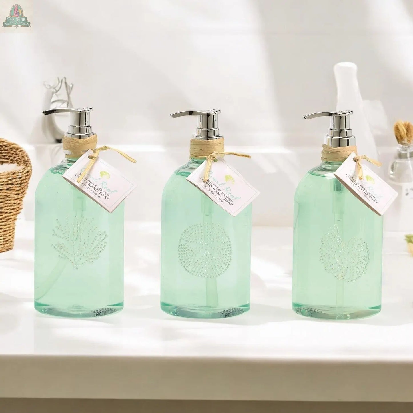 Three By the Sea Hand Soap dispensers with silver pumps, pale green liquid, and etched sea shell designs sit on a white bathroom counter. Each reusable dispenser features a twine-tied tag, perfect for beach-themed bathroom decor.
