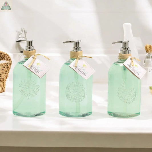 Three By the Sea Hand Soap dispensers with silver pumps, pale green liquid, and etched sea shell designs sit on a white bathroom counter. Each reusable dispenser features a twine-tied tag, perfect for beach-themed bathroom decor.