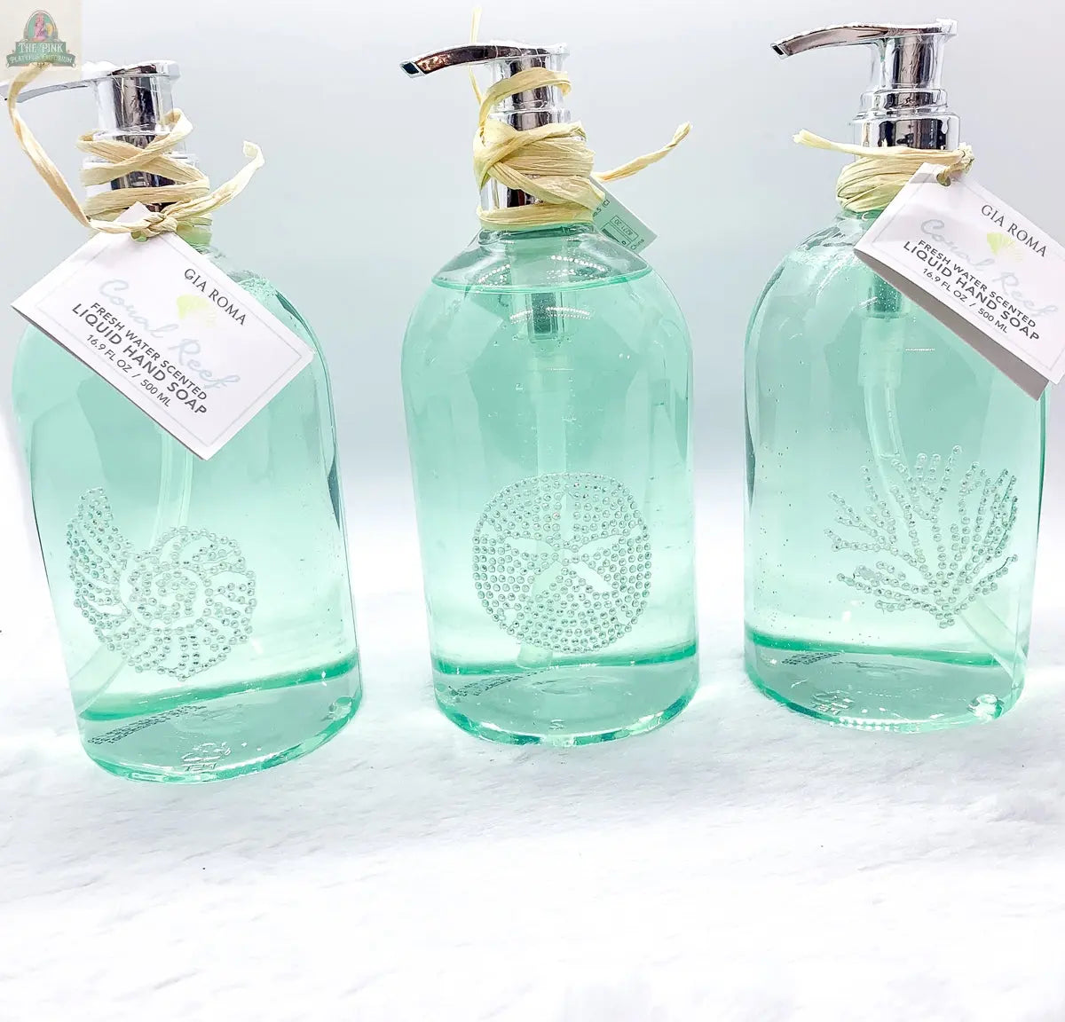 Three clear green By the Sea Hand Soap dispensers with silver pumps, each featuring a coastal white seashell, sand dollar, or coral design plus “Gia Roma” tags and raffia bows—perfect for beach bathroom decor.