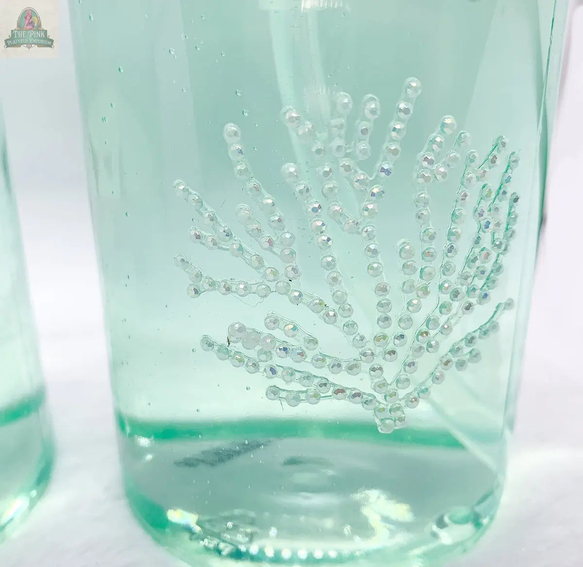 Close-up of By the Sea Hand Soap, a clear green dispenser with a textured, pearl-like coral design. Ideal for beach bathroom decor, its decorative detail and coastal charm stand out against a blurred white background.