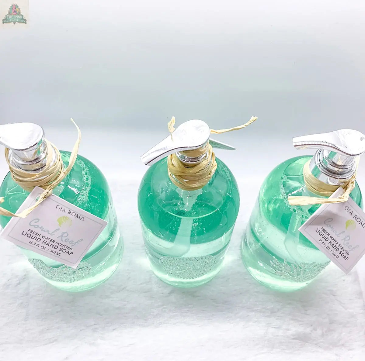 Three clear hand soap dispensers filled with green liquid, each labeled “By the Sea Hand Soap” with raffia ties, are arranged on a white surface—perfect as reusable dispensers or for adding beach-themed bathroom decor.