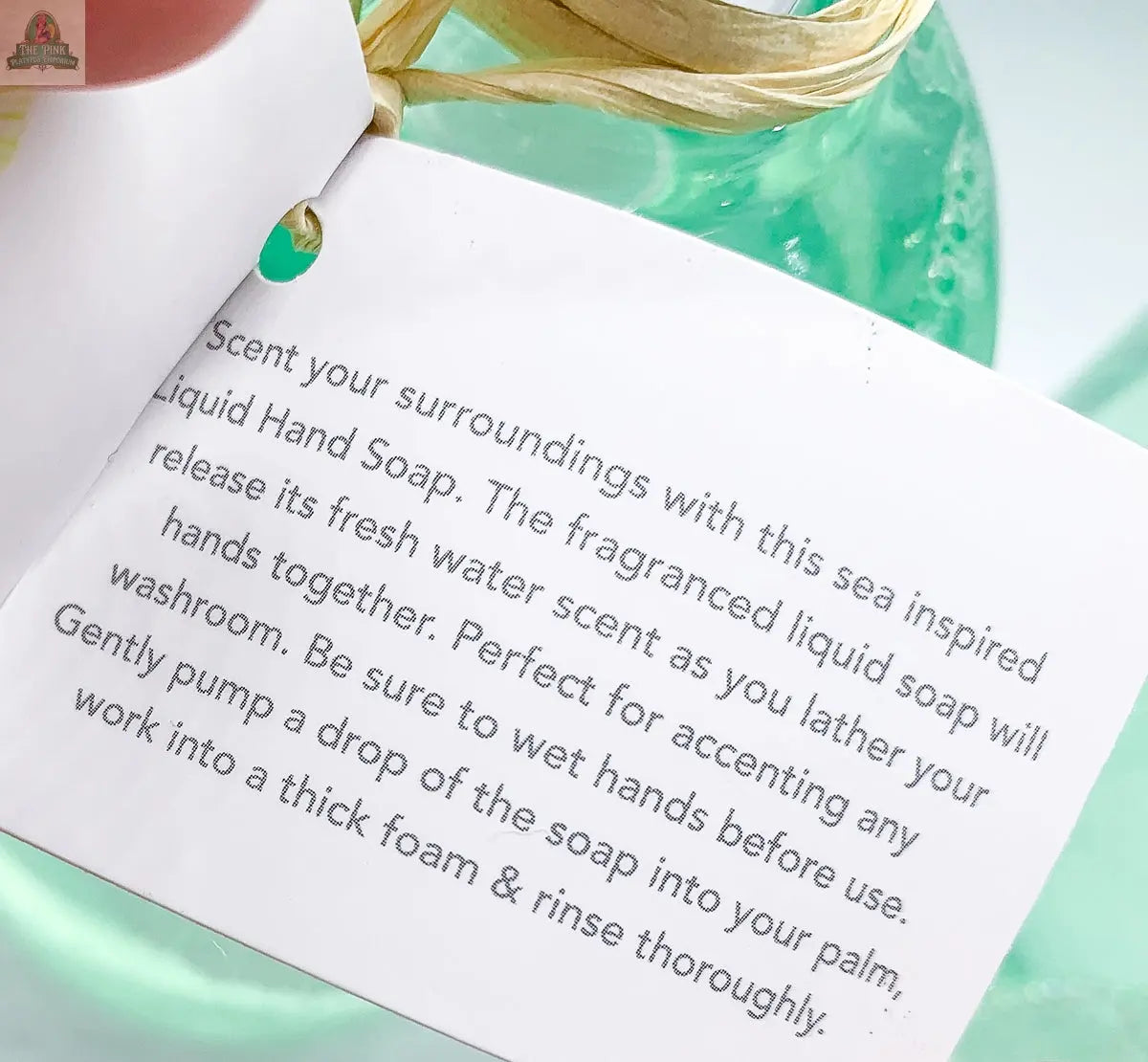 A close-up of the By the Sea Hand Soap label describes its fresh water scent, ideal for beach-themed bathrooms. A green dispenser appears partially behind the label.