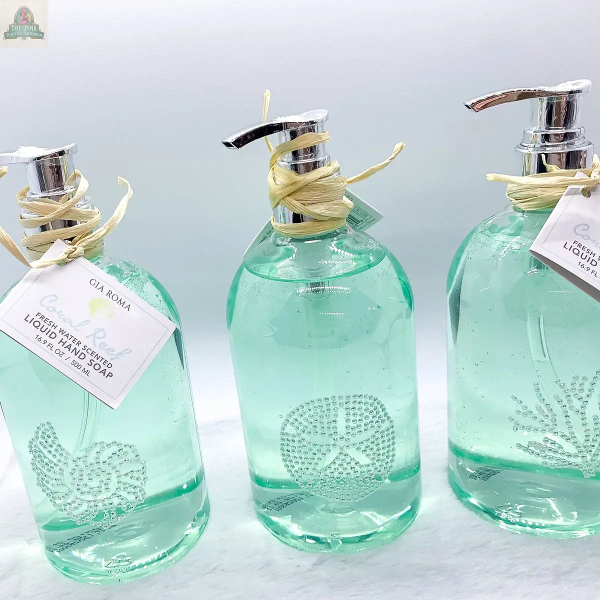 Three By the Sea Hand Soap dispensers filled with light green soap feature sea-themed designs—shell, sand dollar, and coral. Each has raffia ribbon and a labeled tag, ideal for adding a beachy touch to your bathroom decor.
