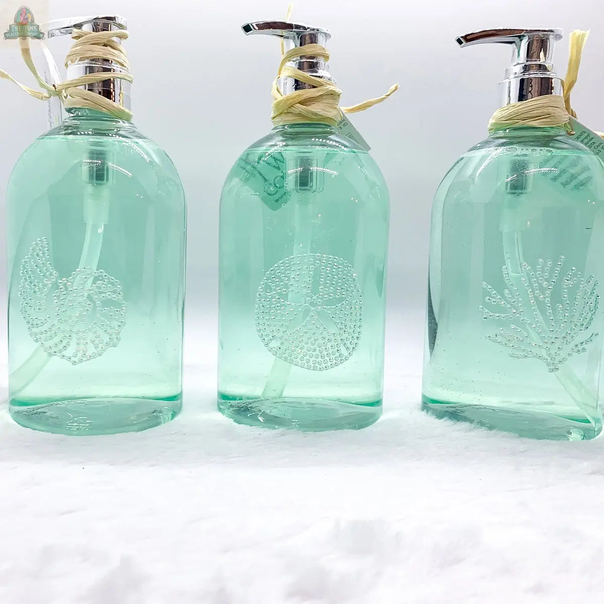 Three green By the Sea Hand Soap dispensers with silver pumps sit in a row on a white surface. Each features a unique white sea-inspired embossed design—nautilus shell, sand dollar, and coral—ideal for beach-themed bathroom decor.