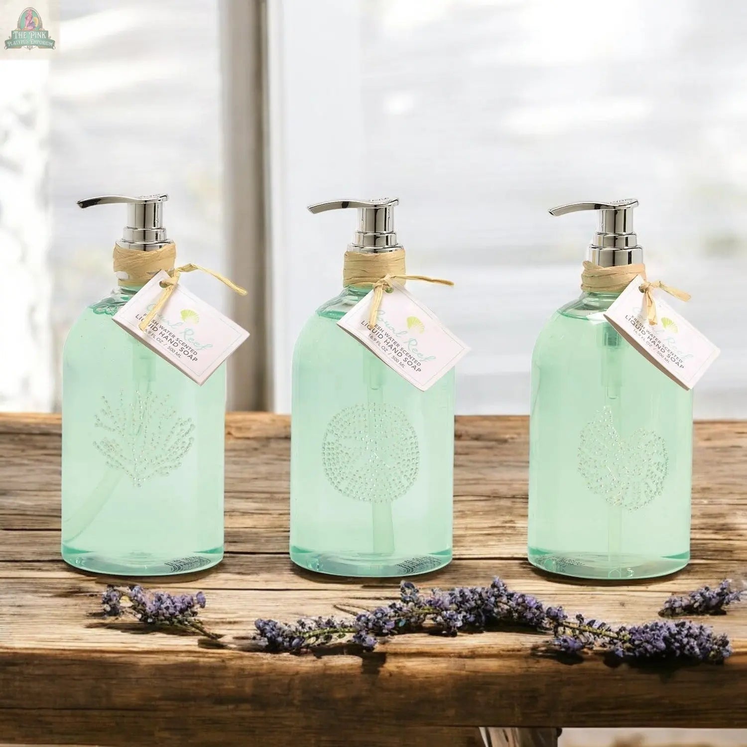Three clear, reusable By the Sea Hand Soap dispensers filled with light blue soap rest on a rustic wood table. Each features an etched seashell, twine-tied tag, and lavender sprigs—perfect for elegant beach-themed bathroom decor.