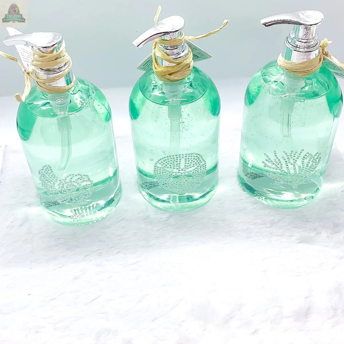 Three By the Sea Hand Soap dispensers—clear glass with silver pumps, filled with light green soap and sea-inspired designs—are arranged on a white textured surface, ideal for beach bathroom decor.