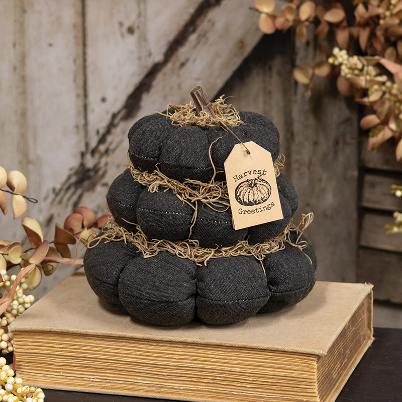 A dark charcoal gray fabric pumpkin decoration wrapped in natural jute twine, resting on a vintage-style book. It features a hanging kraft paper tag that reads 'Harvest Greetings' and is styled with autumn foliage against a rustic wooden background.