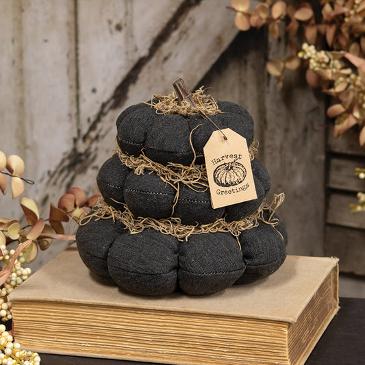 A dark charcoal gray fabric pumpkin decoration wrapped in natural jute twine, resting on a vintage-style book. It features a hanging kraft paper tag that reads 'Harvest Greetings' and is styled with autumn foliage against a rustic wooden background.