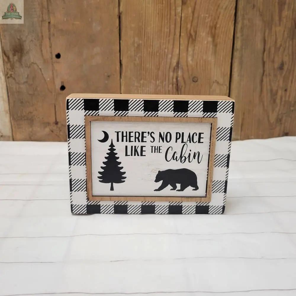 The Cabin Sign features a black and white plaid border and reads "There's No Place Like the Cabin," with pine tree, crescent moon, and bear silhouettes on a wood-style background.