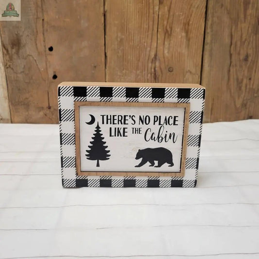 The Cabin Sign features a black and white plaid border and reads "There's No Place Like the Cabin," with pine tree, crescent moon, and bear silhouettes on a wood-style background.