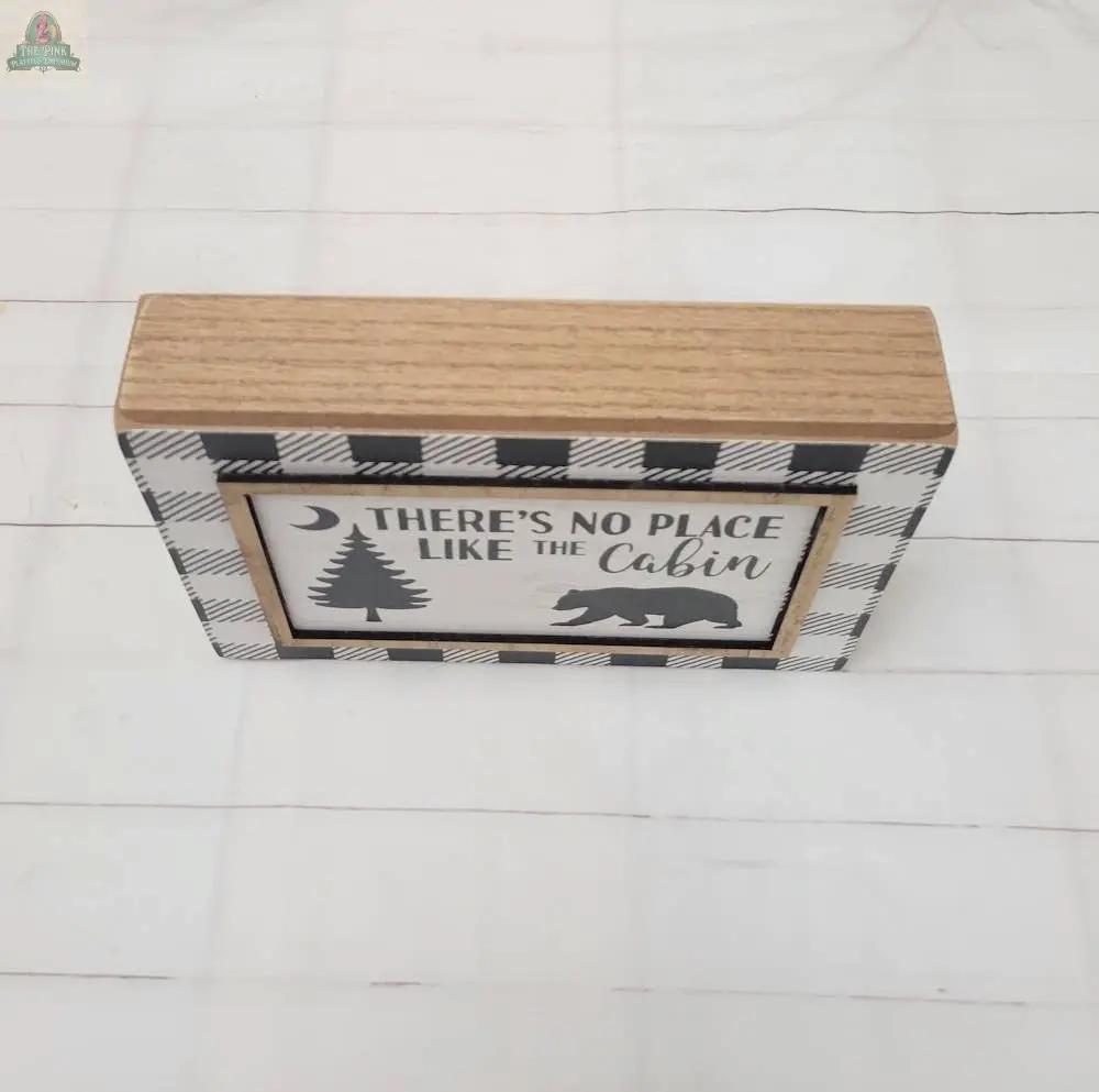 The Cabin Sign features a plaid border, natural wood top, and a white background with silhouettes of a tree, bear, and crescent moon, plus the phrase “There’s no place like the cabin”—a perfect addition to rustic home décor.
