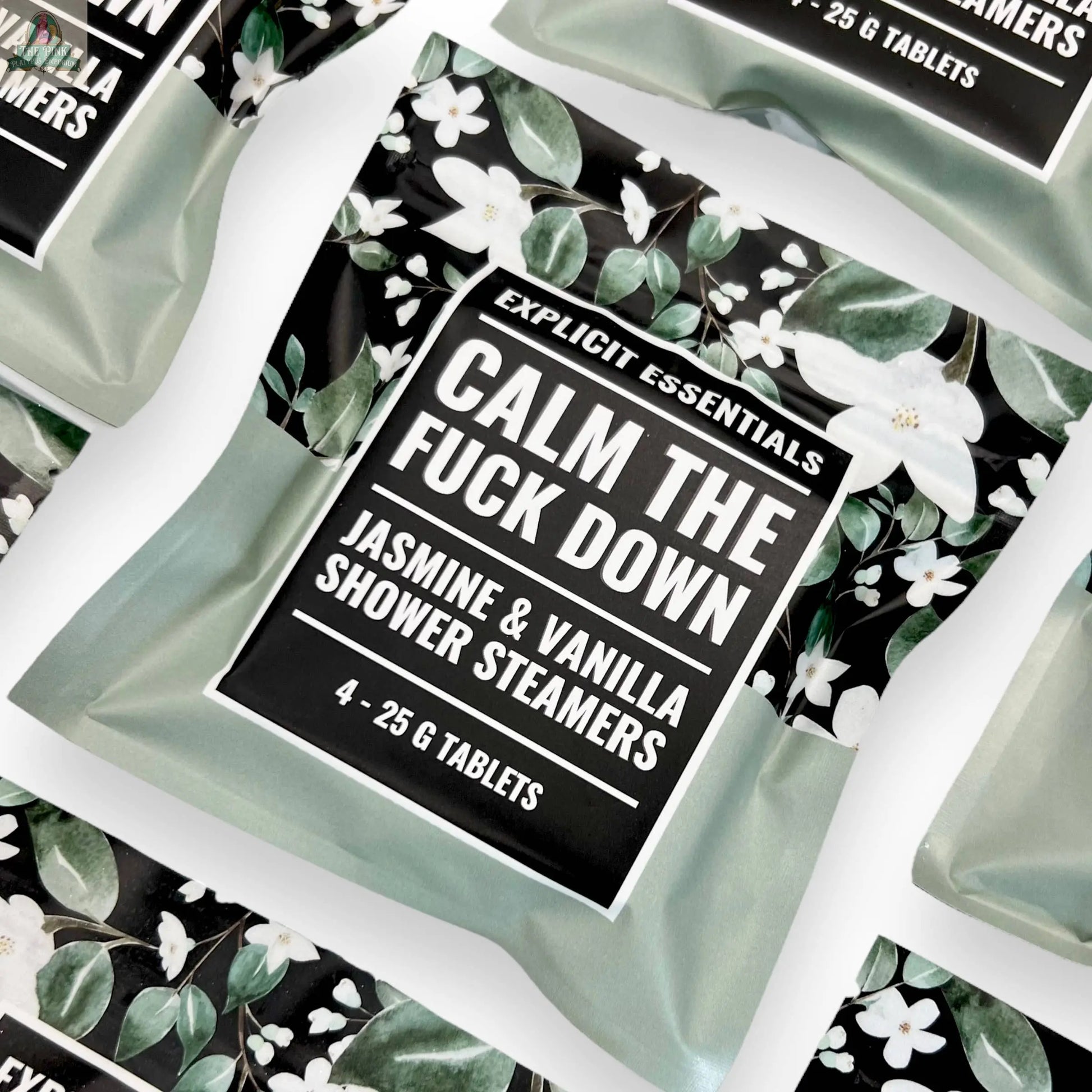 Explicit Essentials Calm TF Down Shower Steamers in Jasmine & Vanilla, with a floral design and bold black-and-white text, are displayed in an appealing flat lay arrangement. Multiple aromatherapy packages are featured.