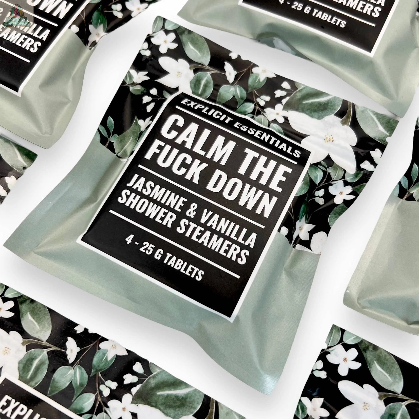 Several sachets of Explicit Essentials Calm TF Down Shower Steamers are arranged on a white surface, featuring jasmine and vanilla scents. The packaging includes floral patterns and bold text for a soothing, stylish look.