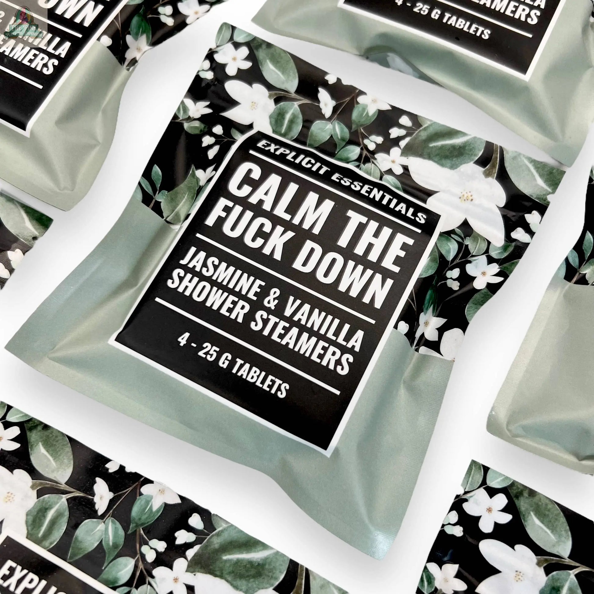 Several sachets of Explicit Essentials Calm TF Down Shower Steamers are arranged on a white surface, featuring jasmine and vanilla scents. The packaging includes floral patterns and bold text for a soothing, stylish look.