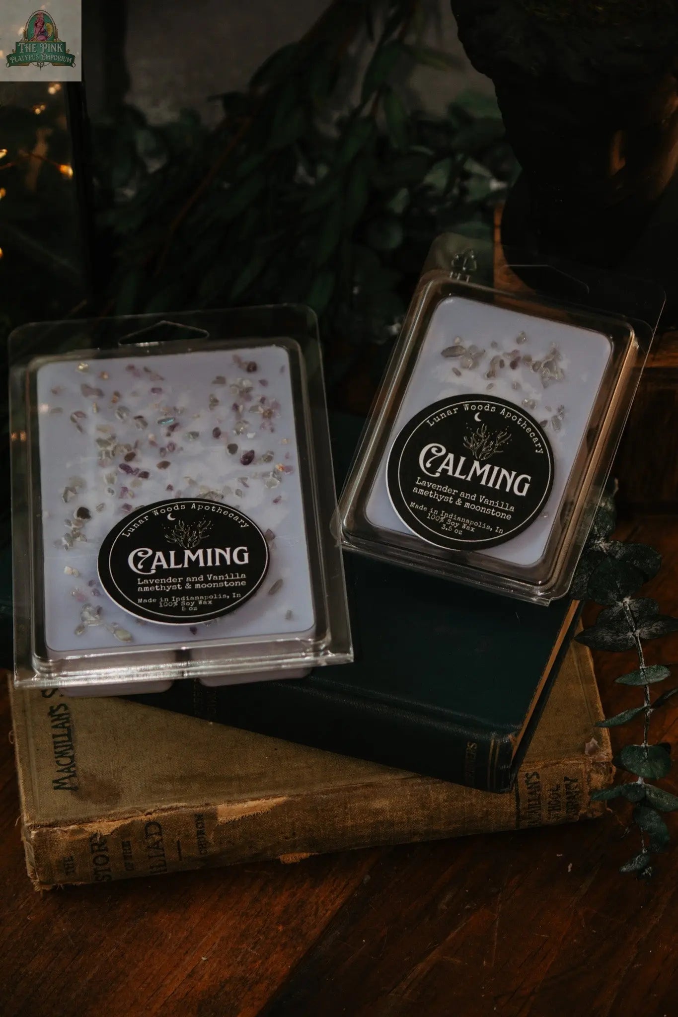 Two packs of Calming — Witchy Wax Melts with dried flowers, set on vintage books and wood, evoke a soothing, weighted blanket scent—ideal for your wax warmer. Greenery in the background adds a peaceful, serene touch.