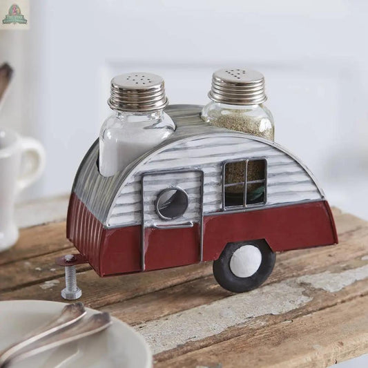 The Camper Salt and Pepper Caddy features glass shakers in a metal holder shaped like a vintage camper trailer, adding charm as a picnic caddy on a rustic wooden table beside plates and cutlery.