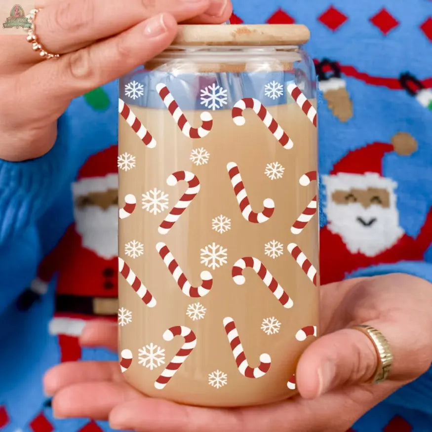 A person holds a Candy Cane holiday glass cup filled with light brown liquid, decorated with red-and-white candy canes and white snowflakes, while wearing a festive blue sweater featuring Santa Claus faces.