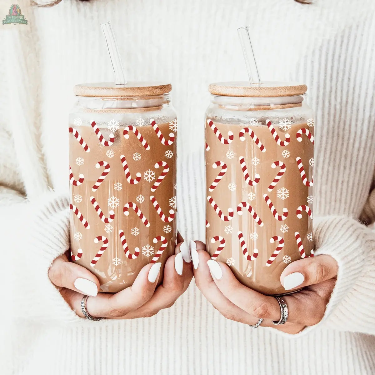 A person in a white sweater holds two iced coffees in Candy Cane holiday glass cups, each topped with a bamboo lid and glass straw. The jars, decorated with candy cane and snowflake designs, are perfect for festive sipping.
