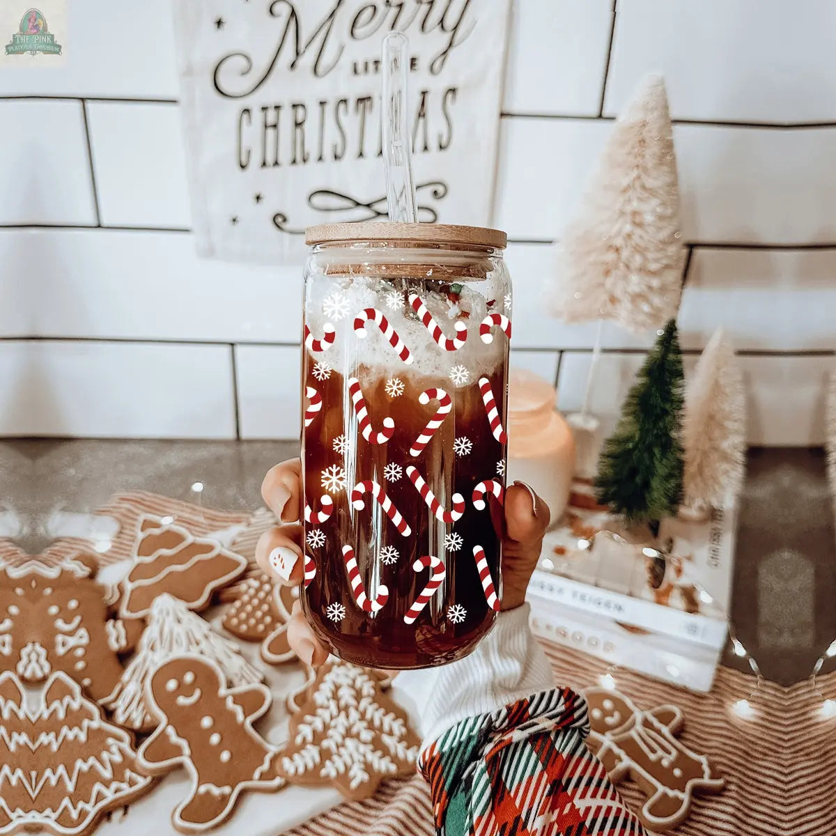 A hand holds a holiday glass of iced coffee topped with whipped cream and Candy Cane sprinkles. Gingerbread cookies, mini trees, and Christmas decorations sit on the table in the background.