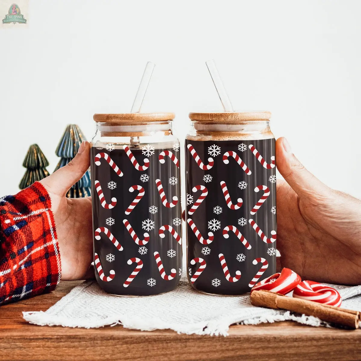 Two hands hold festive glass cups with wooden lids and straws, decorated with candy cane and snowflake patterns. The Candy Cane jars sit near small Christmas tree decorations and candy canes on a wooden surface.