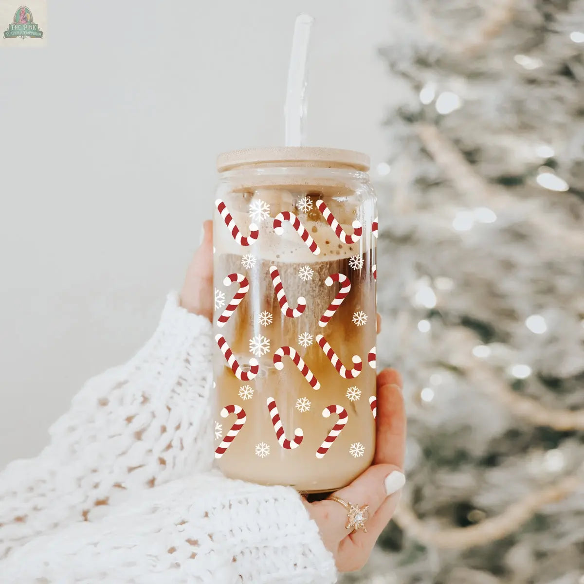 A hand in a white knit sleeve holds a Candy Cane holiday glass cup of iced coffee with a straw, decorated with red candy canes and white snowflakes, in front of a softly glowing Christmas tree in the background.