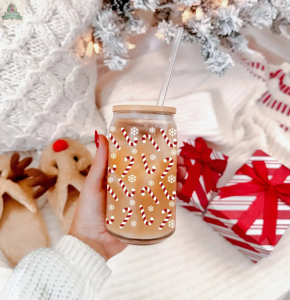 A hand holds a Candy Cane Christmas cup of iced coffee with festive designs, near red and white wrapped gifts, cozy reindeer slippers, and a white knitted blanket with snowy winter decor in the background.