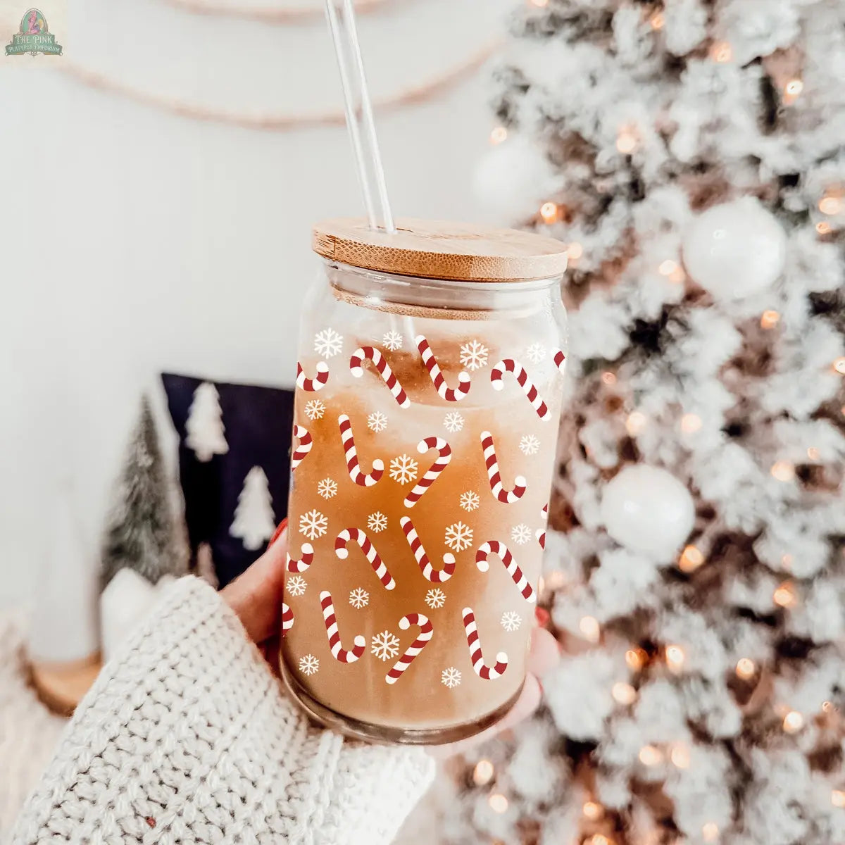 A hand in a cozy knit sweater holds the Candy Cane glass cup with bamboo lid and straw, featuring candy cane and snowflake designs, in front of a white Christmas tree with ornaments.
