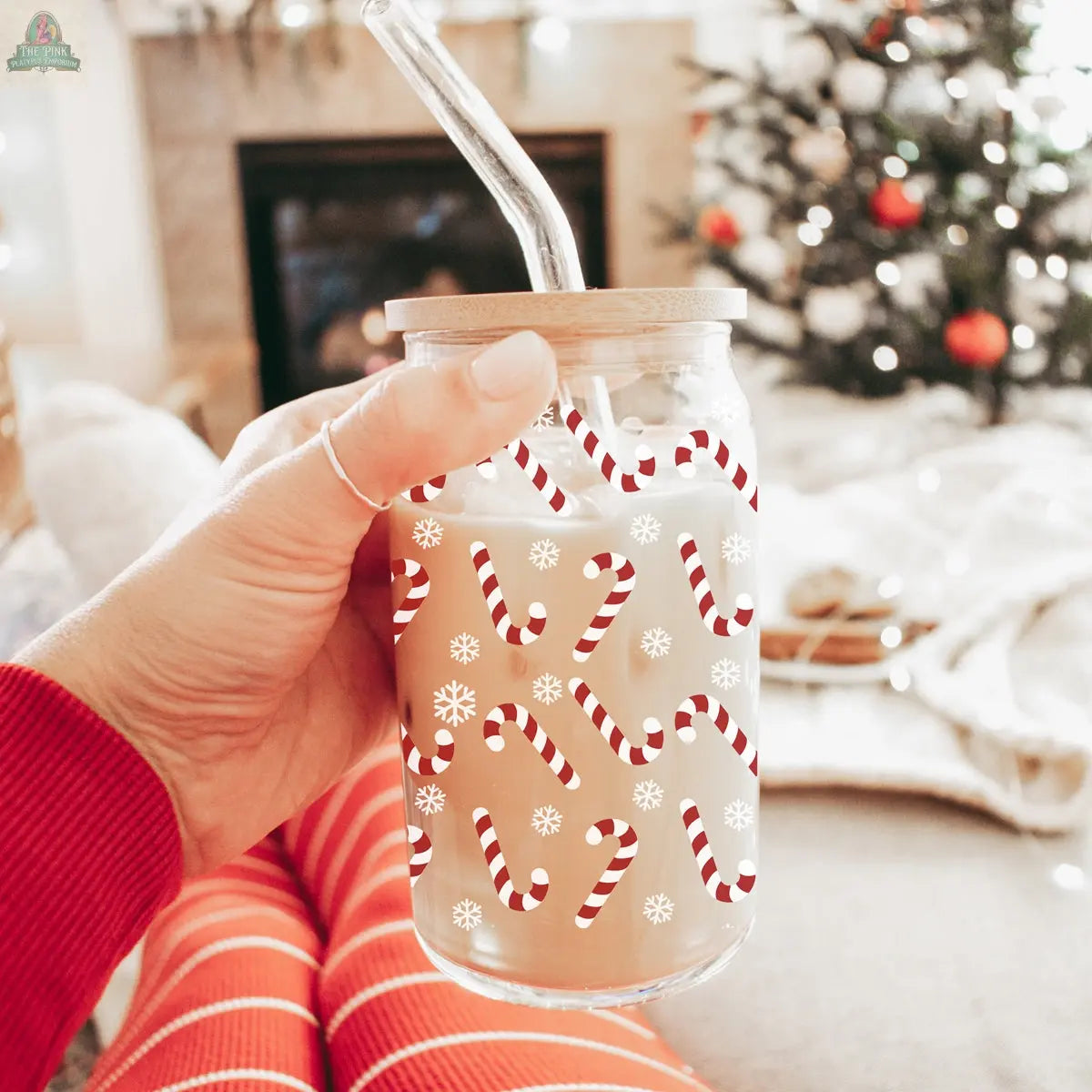 A hand in red pajamas holds the Candy Cane holiday glass cup, featuring a bamboo lid and glass straw. Decorated with candy canes and snowflakes, the festive cup stands out as a blurred ornamented tree glows behind.