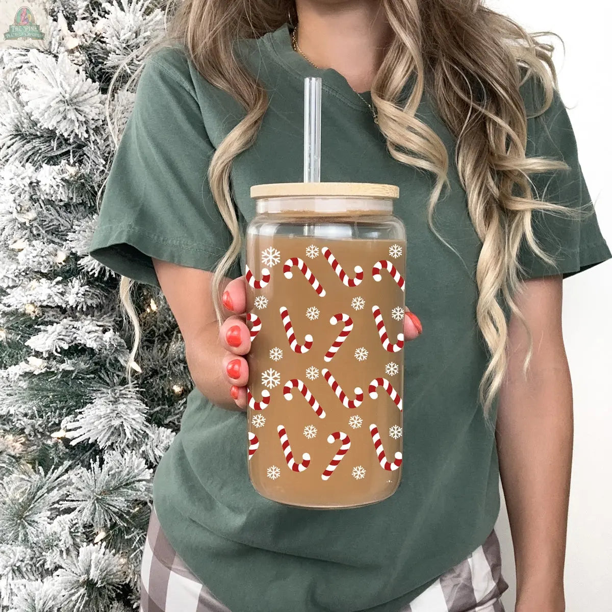 A woman with long wavy hair holds a Candy Cane holiday glass cup, decorated with festive candy canes and snowflakes, filled with a light brown drink, while standing before a beautifully frosted Christmas tree.