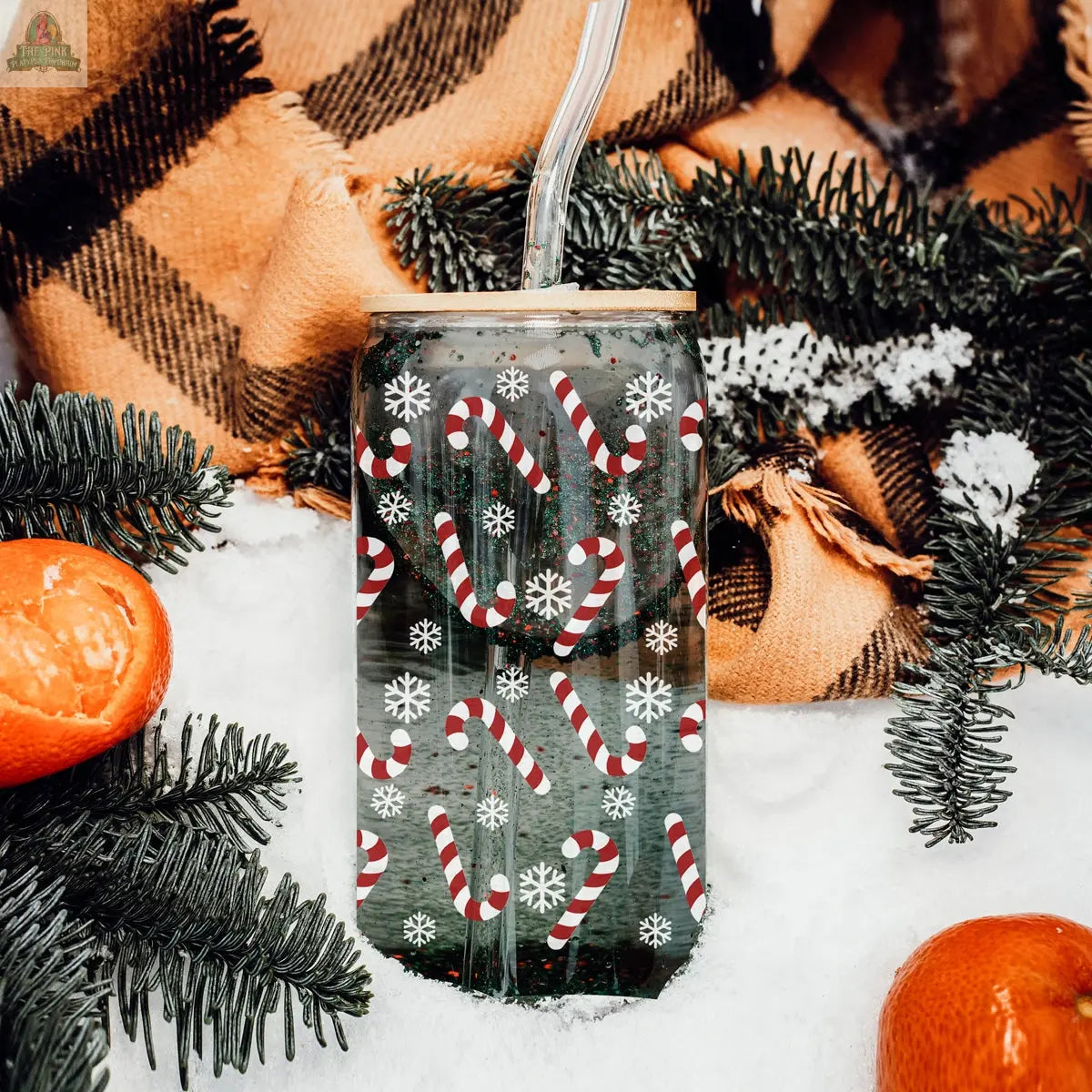 The Candy Cane holiday glass cup with a clear straw, decorated with candy canes and snowflakes, rests on snow among pine branches, tangerines, and a brown plaid blanket.