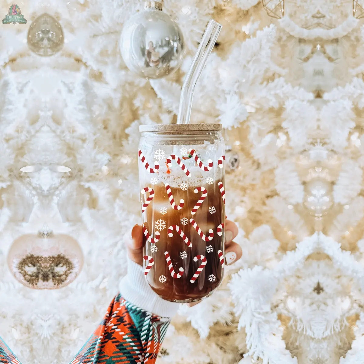 A hand holds a festive Candy Cane iced coffee cup with a clear straw, featuring candy cane and snowflake designs. In the background, a white Christmas tree decorated with silver ornaments is visible.