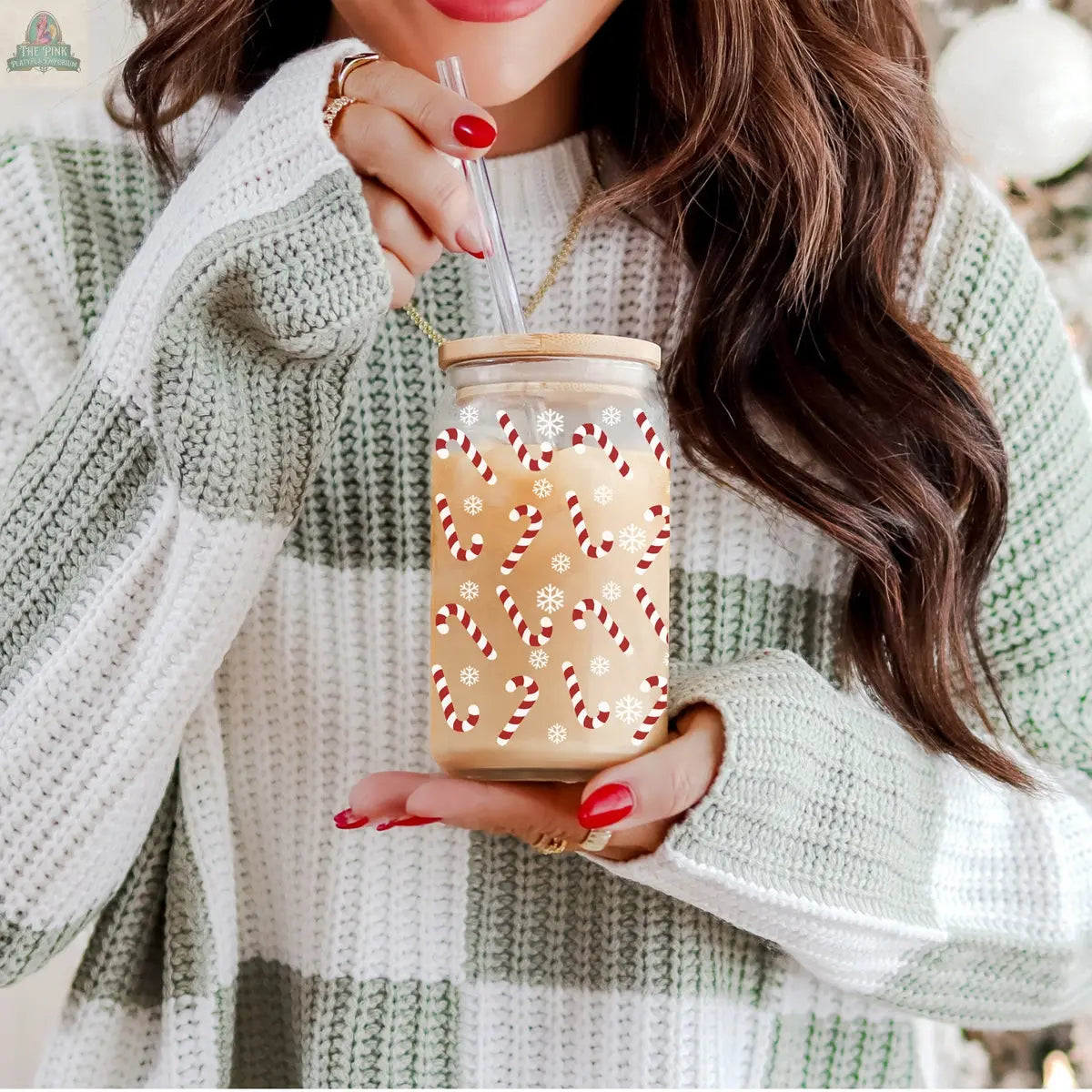 A woman with long brown hair and red nails holds the Candy Cane Christmas cup, featuring candy cane and snowflake designs. She wears a green and white striped sweater.
