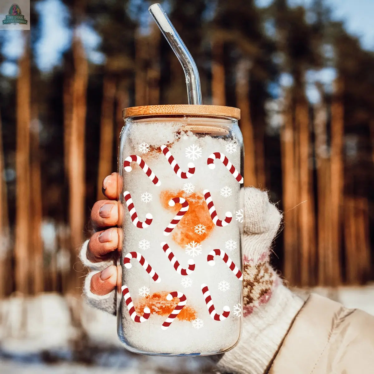 A gloved hand holds the Candy Cane Christmas mason jar with a bamboo lid and metal straw, filled with snow. The jar features candy cane and snowflake designs, set against pine trees and a snowy background.