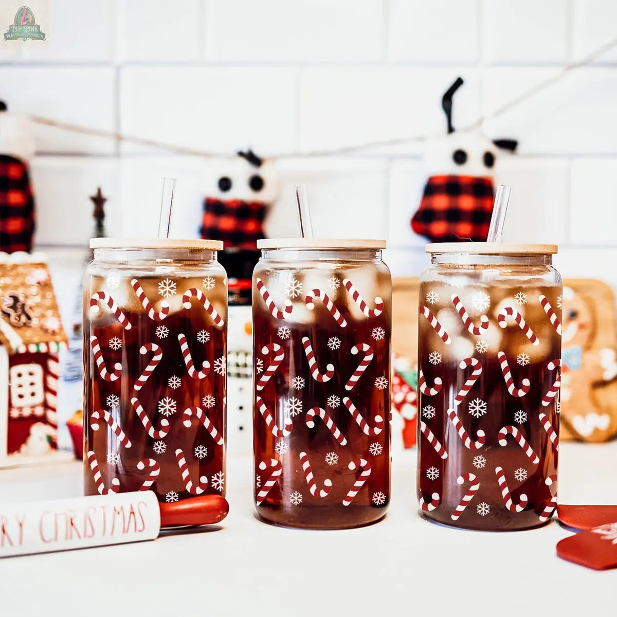 Three iced Candy Cane drinks in clear holiday glass cups with candy cane and snowflake designs, each with a straw. Christmas stockings and festive decorations appear in the background for a holiday-themed scene.