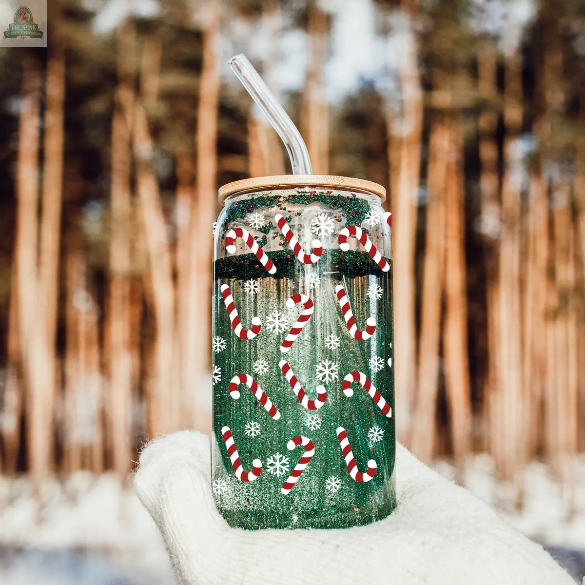 A gloved hand holds the Candy Cane cup with a bamboo lid and glass straw, featuring candy cane and snowflake designs. Blurred pine trees and snow are in the background.