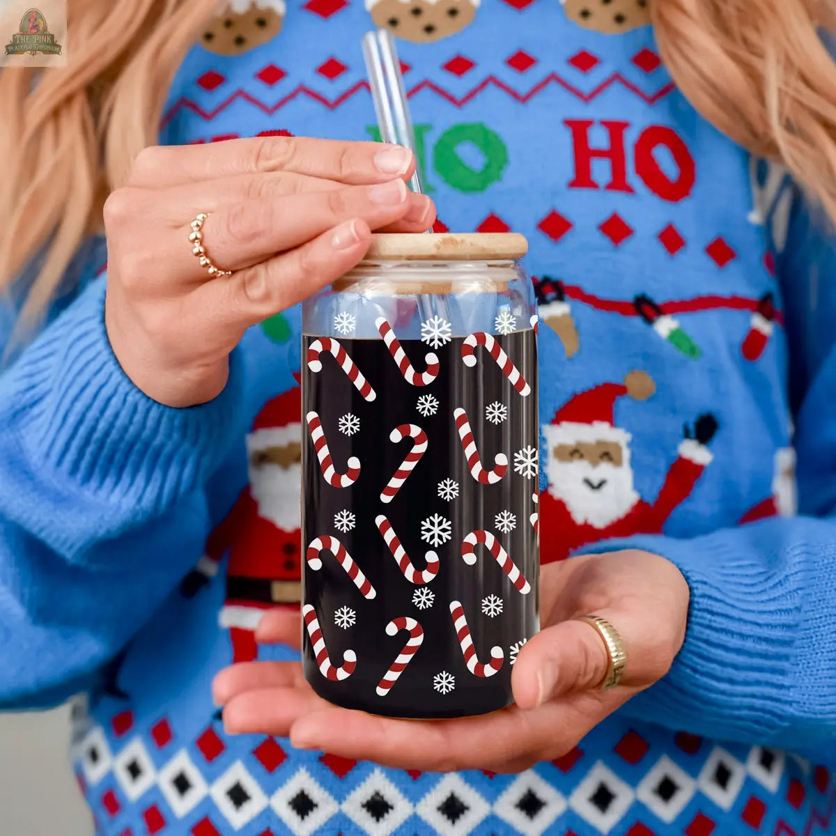 A person wearing a festive blue Christmas sweater with Santa and “HO HO HO” text holds a Candy Cane holiday glass cup with a straw, decorated with candy canes and snowflakes.