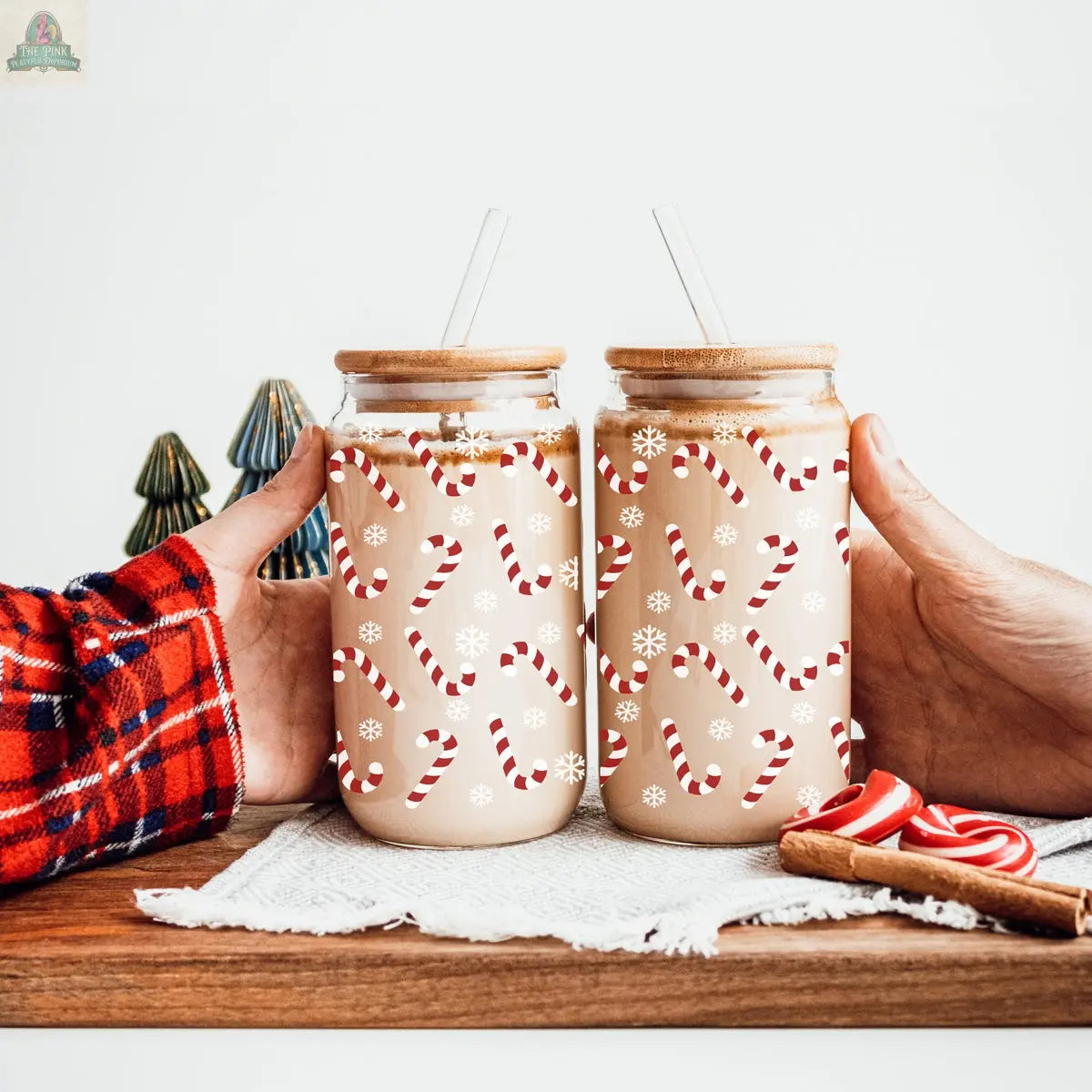 Two hands hold festive Christmas cups with candy cane and snowflake designs, filled with a creamy drink and topped with wooden lids and straws. A Candy Cane product rests on a wooden tray beside small ceramic trees.