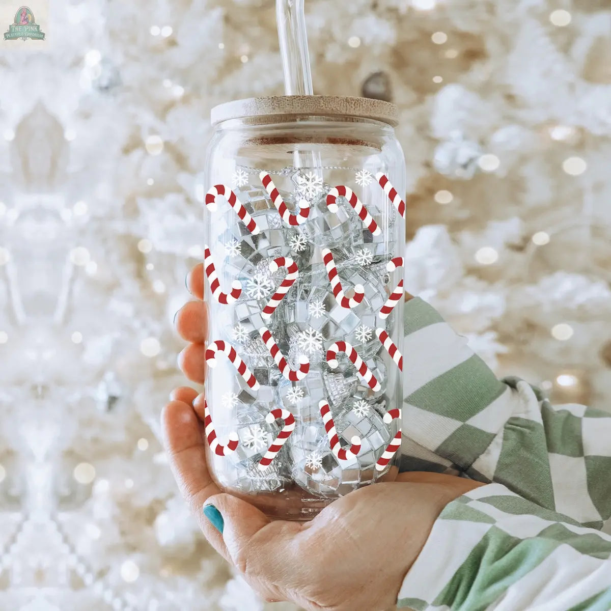A person in a green and white plaid shirt holds the Candy Cane holiday glass cup with bamboo lid and straw, featuring candy cane and snowflake designs. A blurred white Christmas tree is seen in the background.