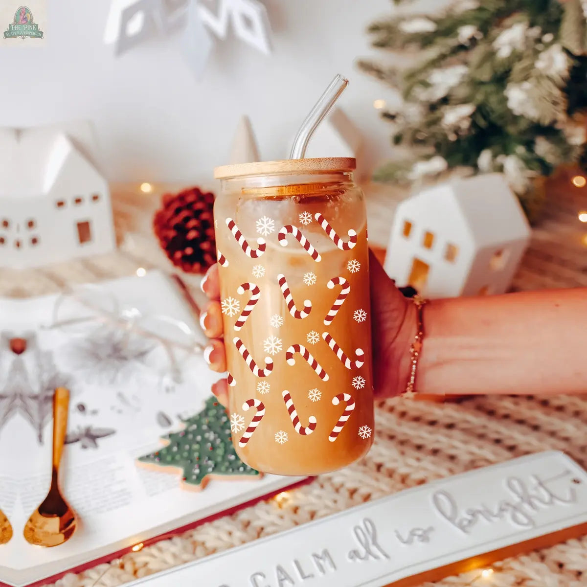 A hand holds the Candy Cane holiday glass cup with a bamboo lid and glass straw, filled with a light brown drink. The Christmas cup, decorated with candy cane and snowflake patterns, sits amid festive décor and a book in the background.