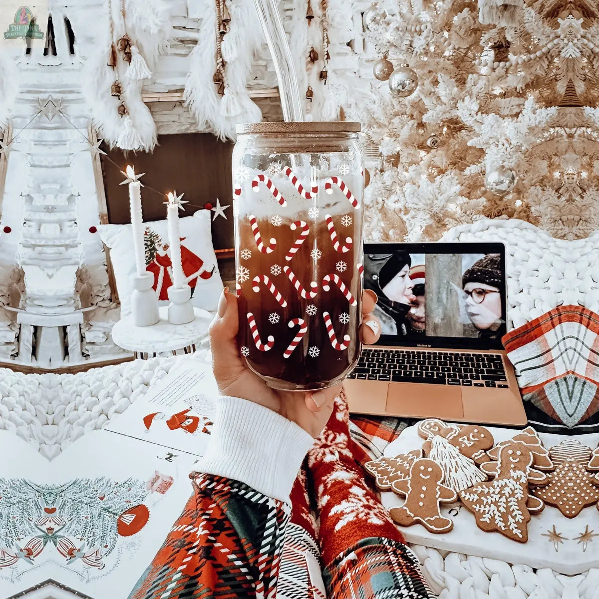 A hand holds a Candy Cane Christmas cup filled with a festive drink in a cozy, holiday-themed setting. Nearby are gingerbread cookies, a laptop playing a movie, and white and red decorations.