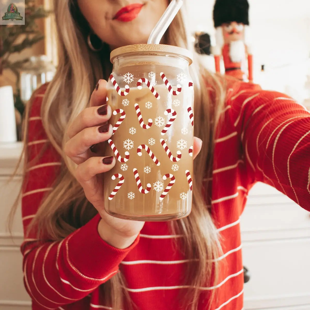 A woman in a red striped sweater holds the Candy Cane holiday glass cup with candy cane and snowflake designs, filled with a light brown drink and reusable straw. A nutcracker is visible in the background.
