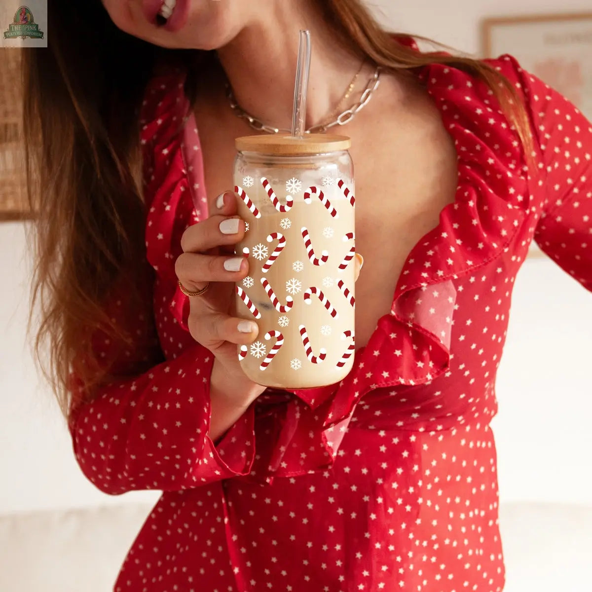 A smiling woman in a red, star-patterned dress holds the “Candy Cane” Christmas mason jar with straw, decorated with candy cane and snowflake designs and filled with a creamy drink. Her head is partly out of frame.