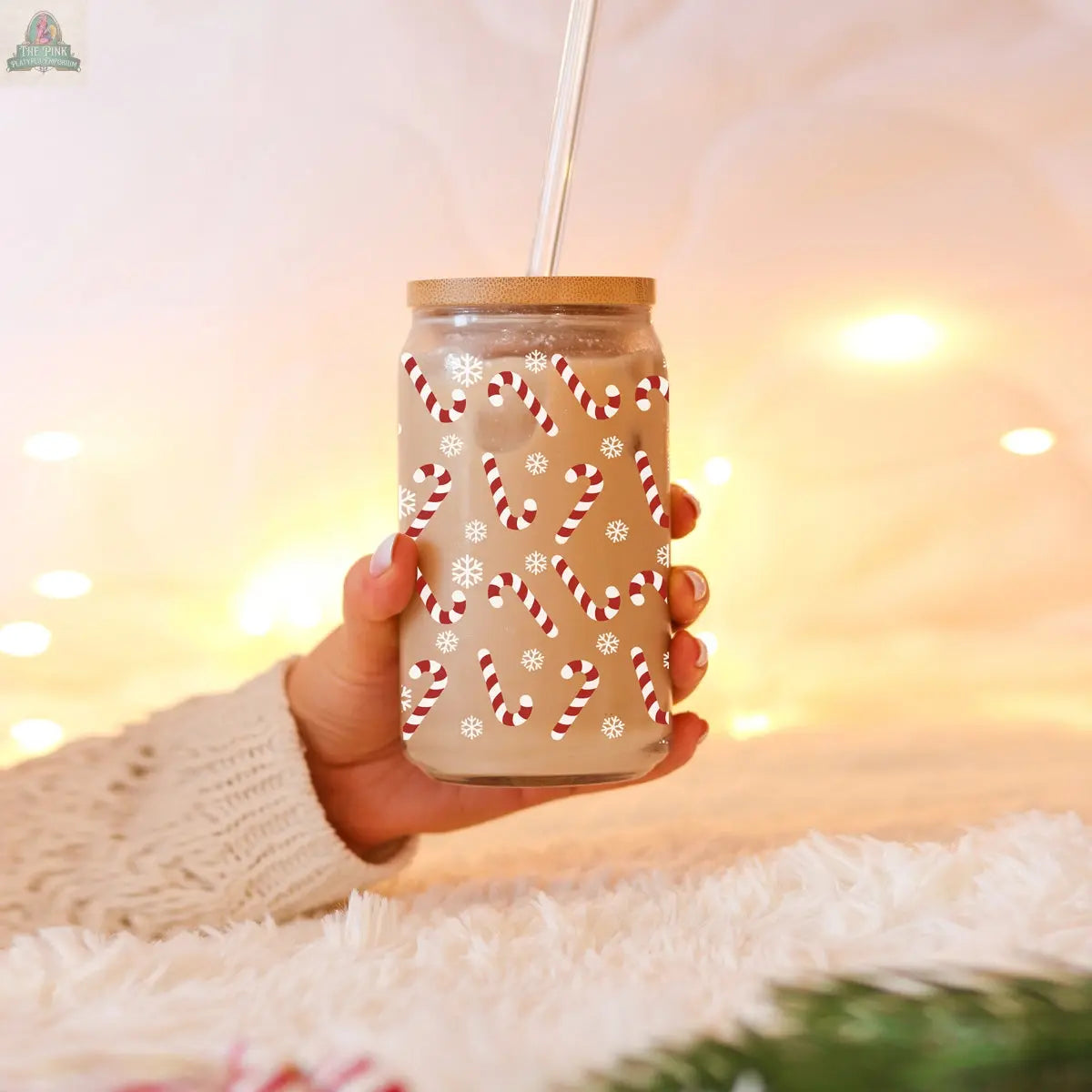 A hand in a cozy sweater holds the Candy Cane holiday glass cup with lid and straw, featuring red and white candy canes and snowflakes. Warm yellow lights blur in the background, adding to the festive mood.