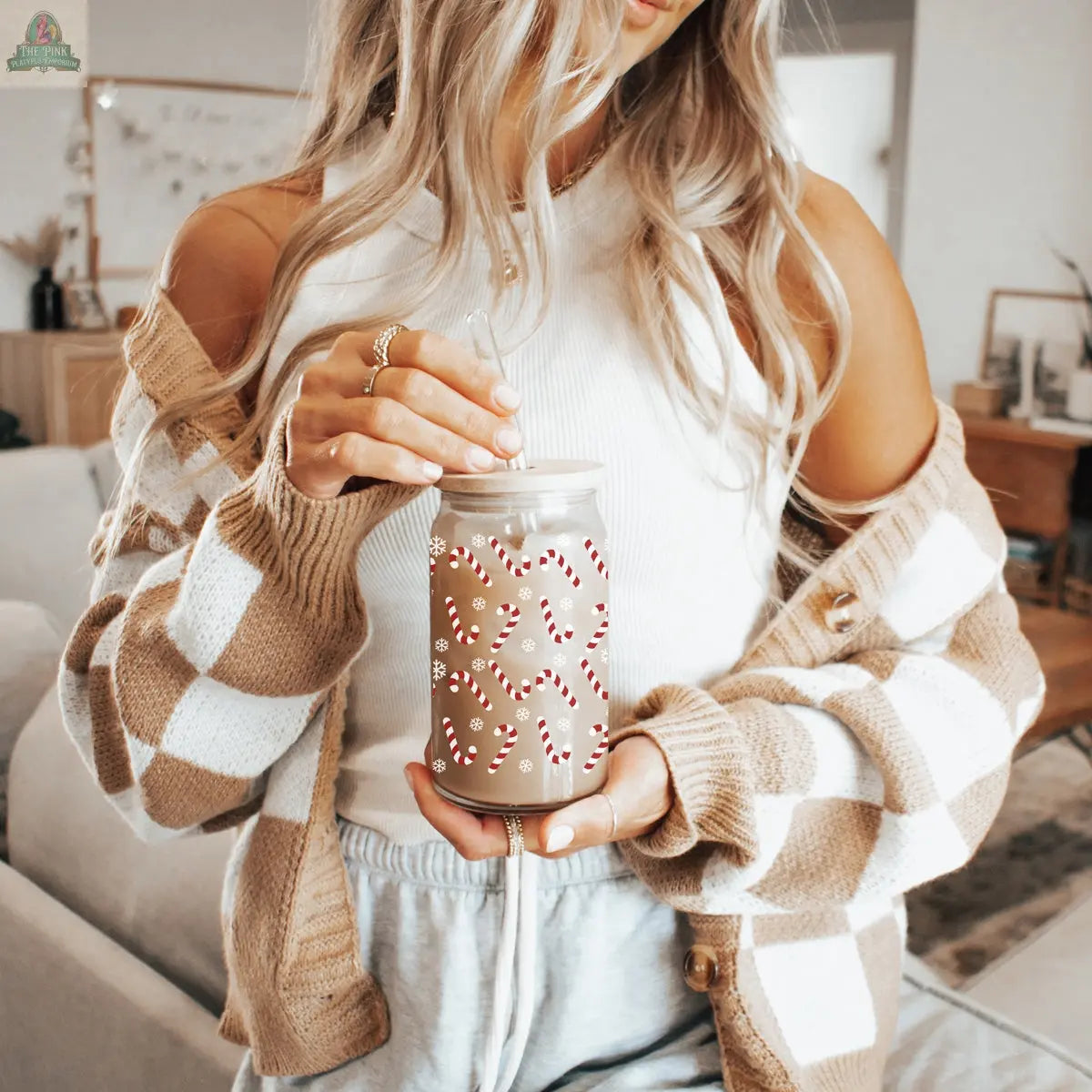 A woman with long, wavy blonde hair stands in a cozy living room, holding a festive cup featuring the Candy Cane product. She wears a white tank top, a tan and white checkered cardigan, and gray lounge pants.