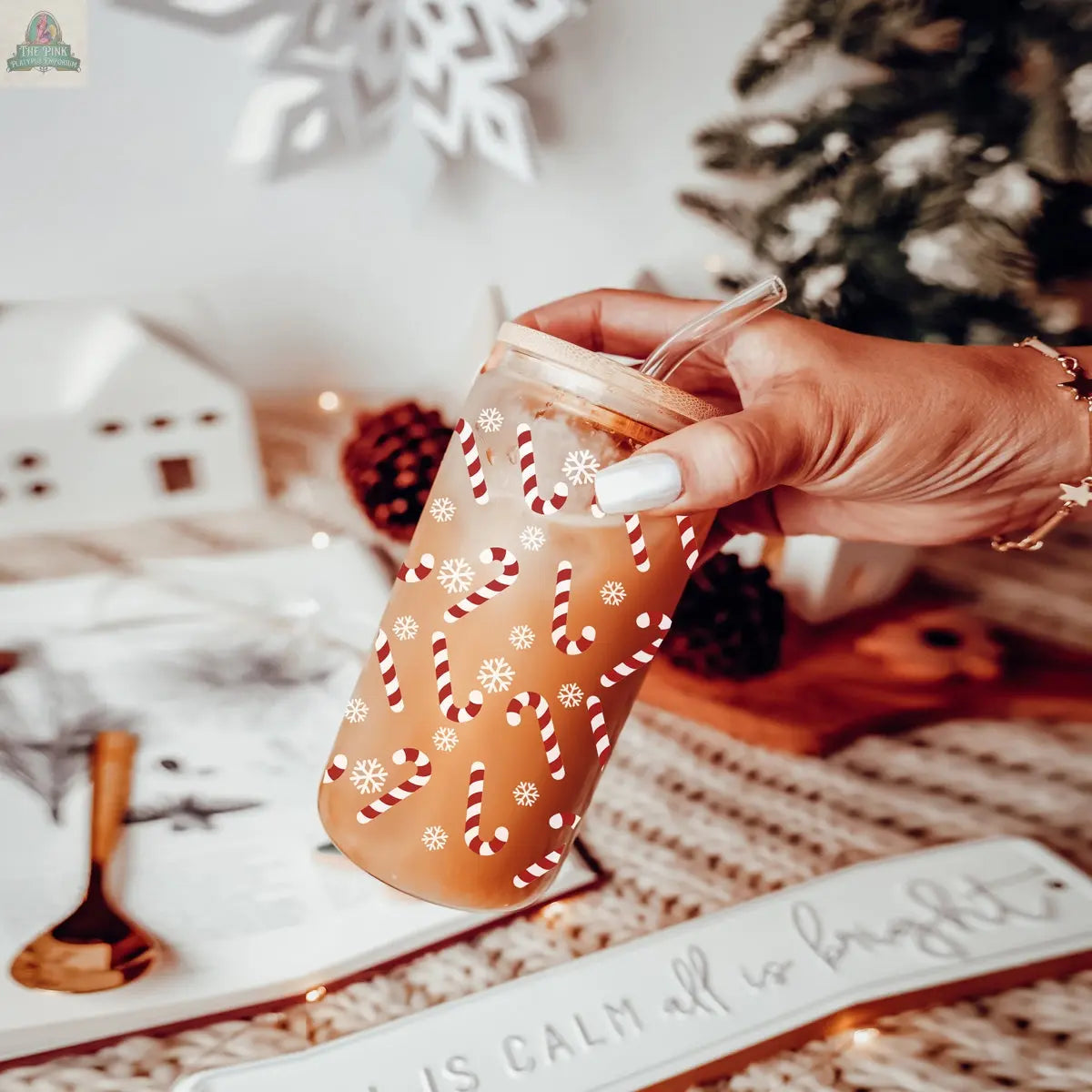 A hand holds an iced coffee in a Candy Cane Christmas cup decorated with candy canes and snowflakes. Holiday-themed items—a snowflake ornament, pinecone, mini house, and small tree—are displayed in the background.