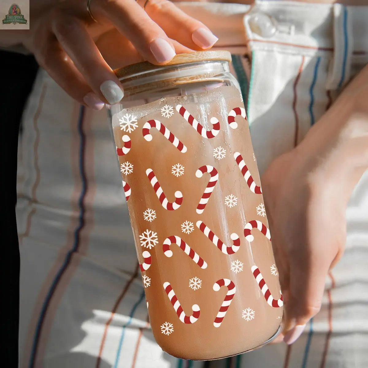 A person in striped pants holds a Candy Cane holiday glass cup decorated with red and white candy canes and white snowflakes, filled with a light brown beverage.